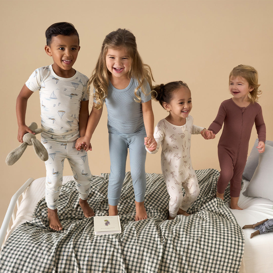 Four happy kids in cozy pajamas jumping on a bed together. Click here to discover cozy sleep styles from Modern Moments.